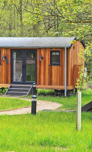 Cabane glamping en bois : Escapade nature et confort Cabane de glamping en bois, toit ondulé gris. Double porte et fenêtre sombres. Sentier de gravier, pelouse verte et forêt au printemps.